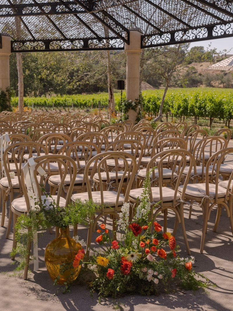 Rows of bentwood ceremony chairs under an iron pergola with an orange and coral floral arrangement