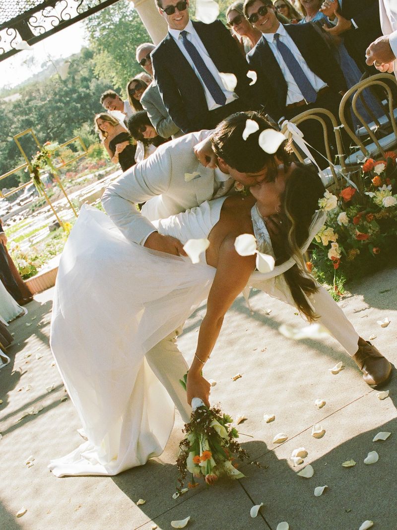 Newlyweds share a kiss at the end of the aisle as guests toss petals at an outdoor vineyard ceremony