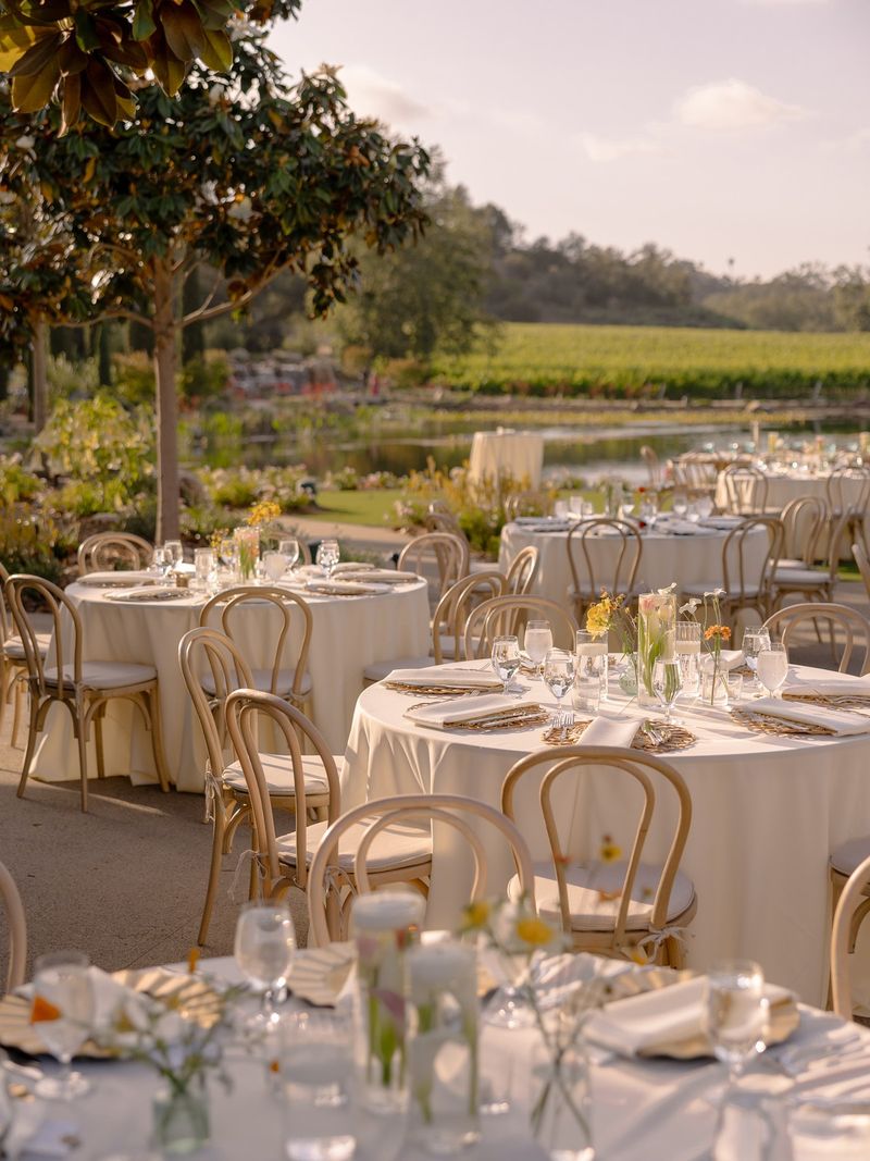 Round reception tables with cream linens and bentwood chairs at golden hour beside a vineyard pond