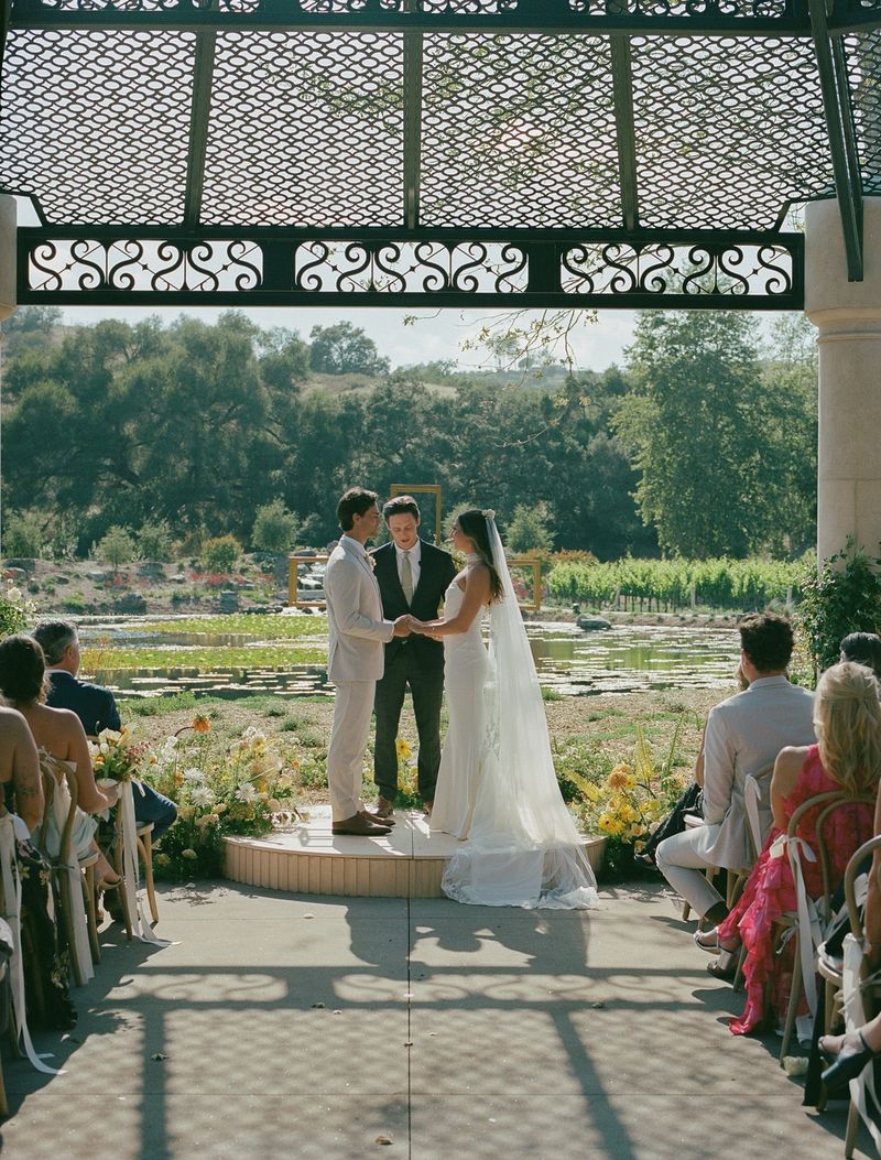Couple exchanging vows beneath an ornate iron pergola with vineyards and a pond beyond