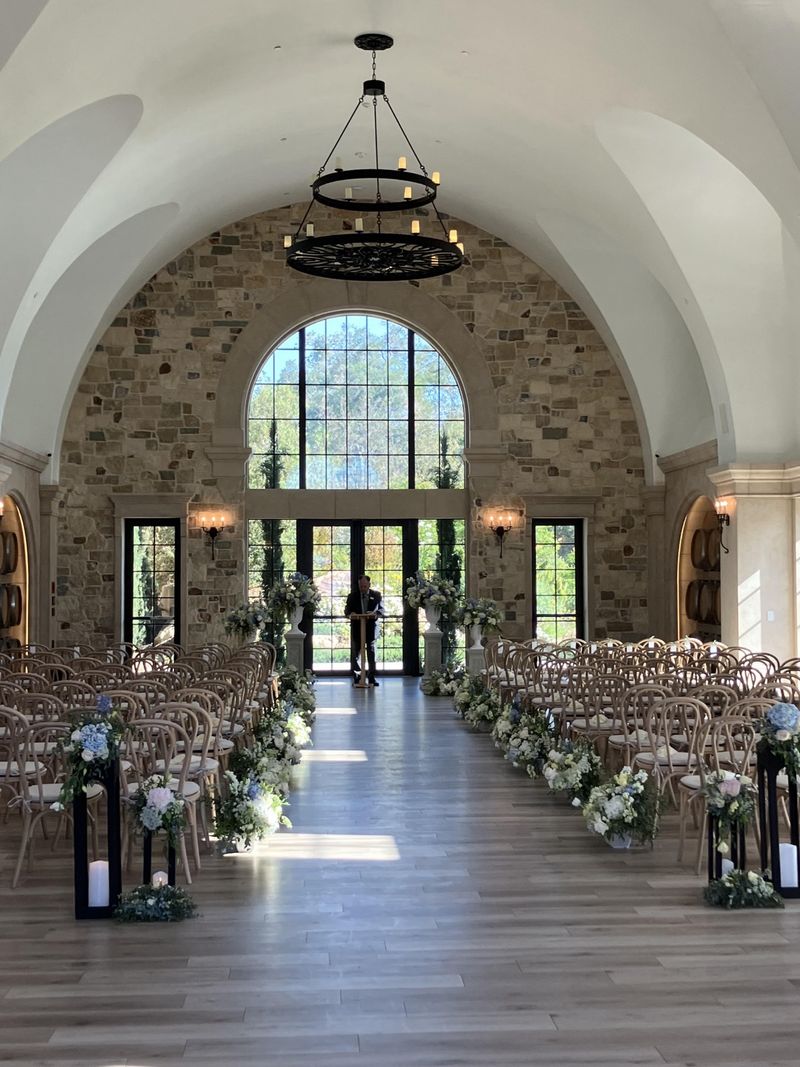 Ceremony hall with a stone arch, iron chandelier and chairs lining an aisle decorated with white and blue florals